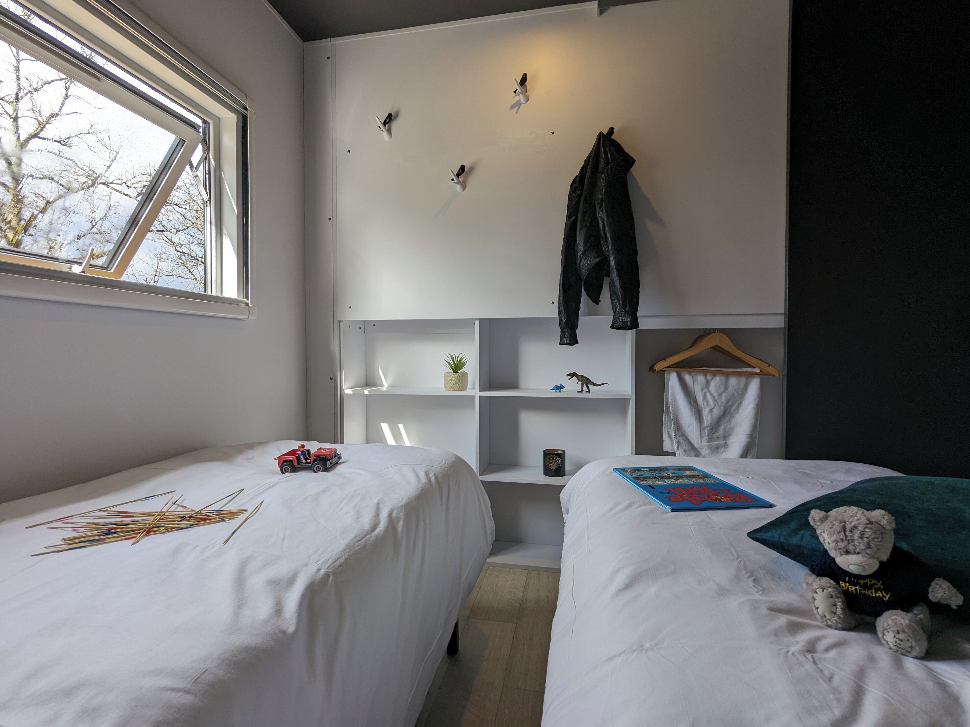 Un dormitorio con dos camas, una ventana, una repisa con una planta, un osito de peluche y una chaqueta colgada en la pared.