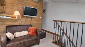 A living room with a brown leather sofa, a flat-screen TV mounted on a wooden wall, a small table with a lamp, and a staircase with a wooden railing.