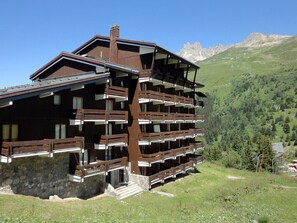 A multi-story wooden chalet with balconies, set against a mountainous backdrop.