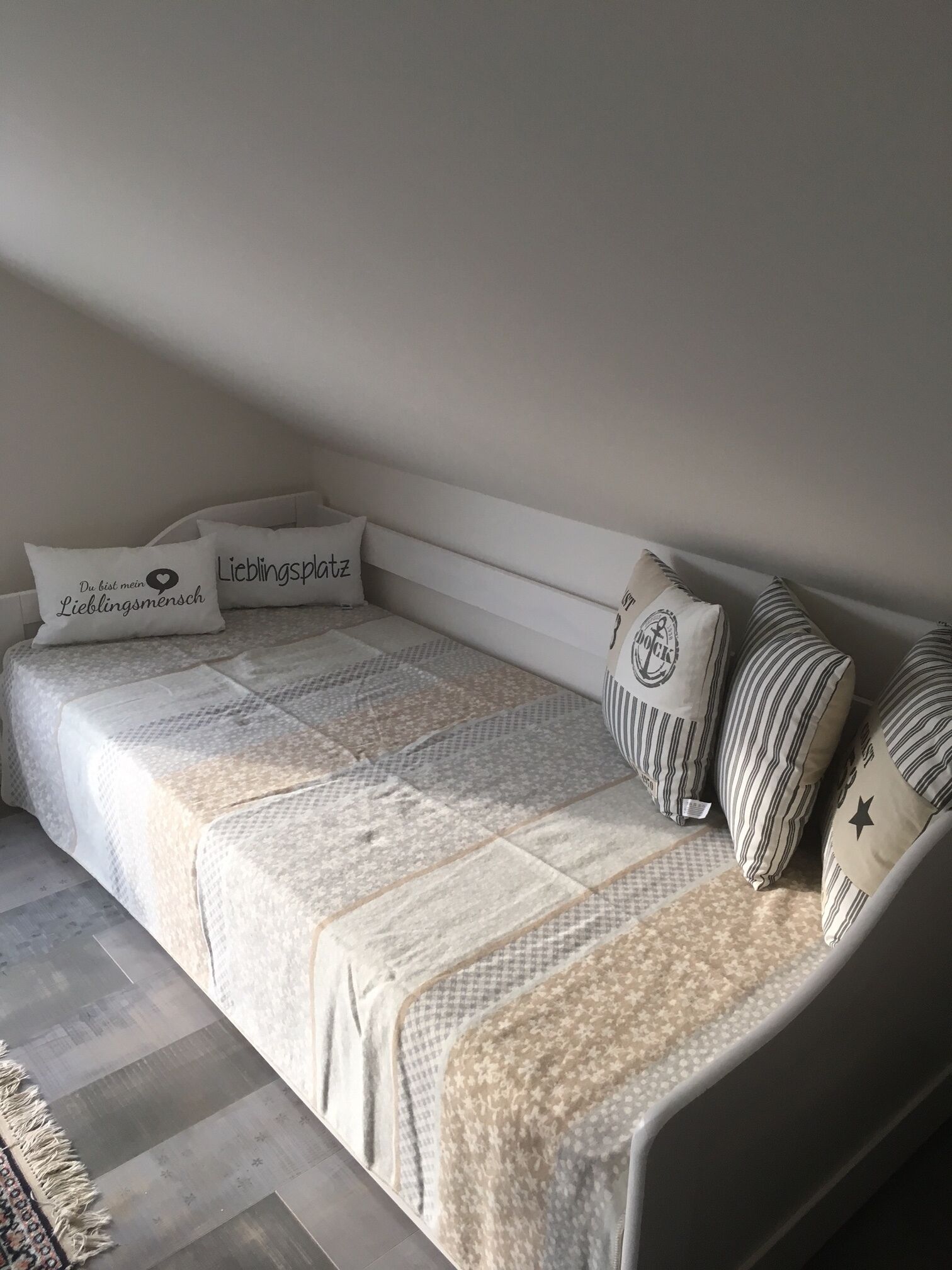 A single bed with a patterned bedspread and pillows, a striped pillow, and a small rug on a wooden floor.