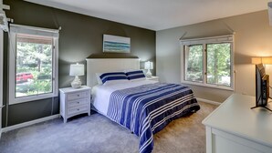 A bedroom with a bed, two bedside tables, a nightstand, a desk, and two windows.