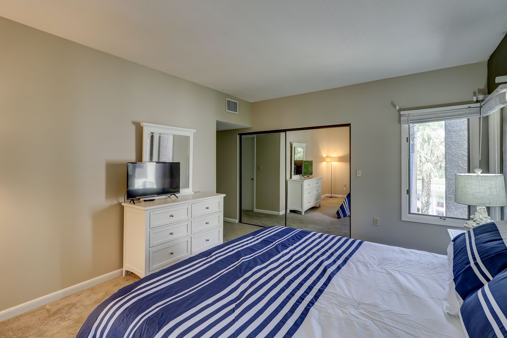 A bedroom with a bed, a dresser, a TV, a mirror, and a window.