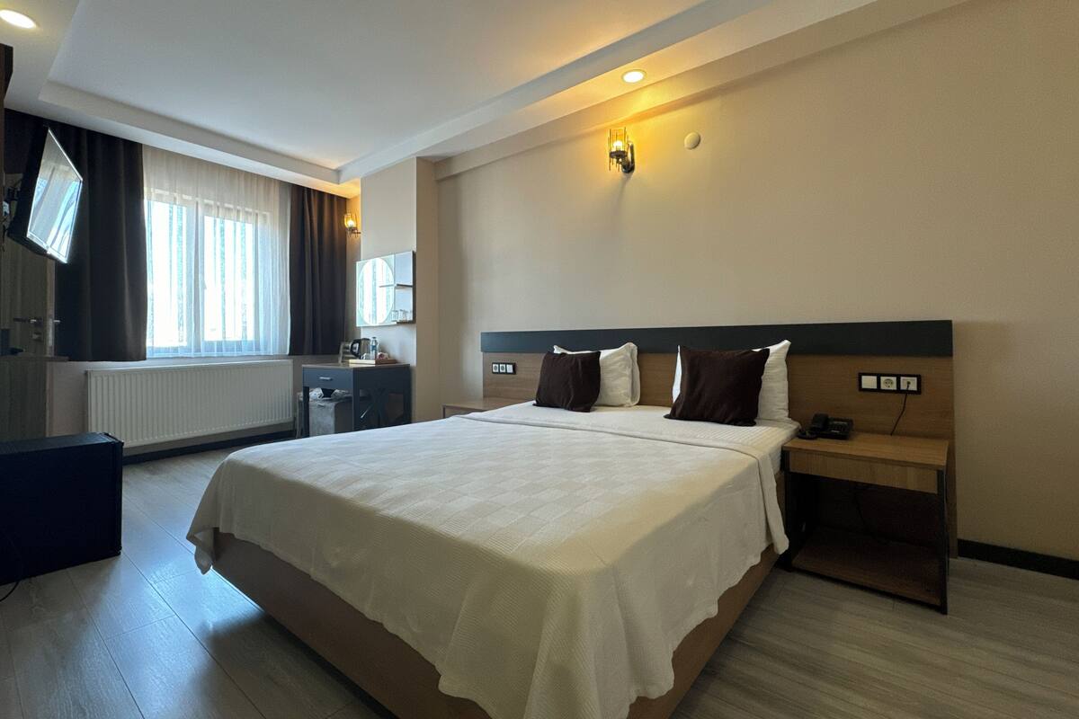 A hotel room with a large bed, bedside tables, a desk, a chair, a mirror, and a window with curtains.