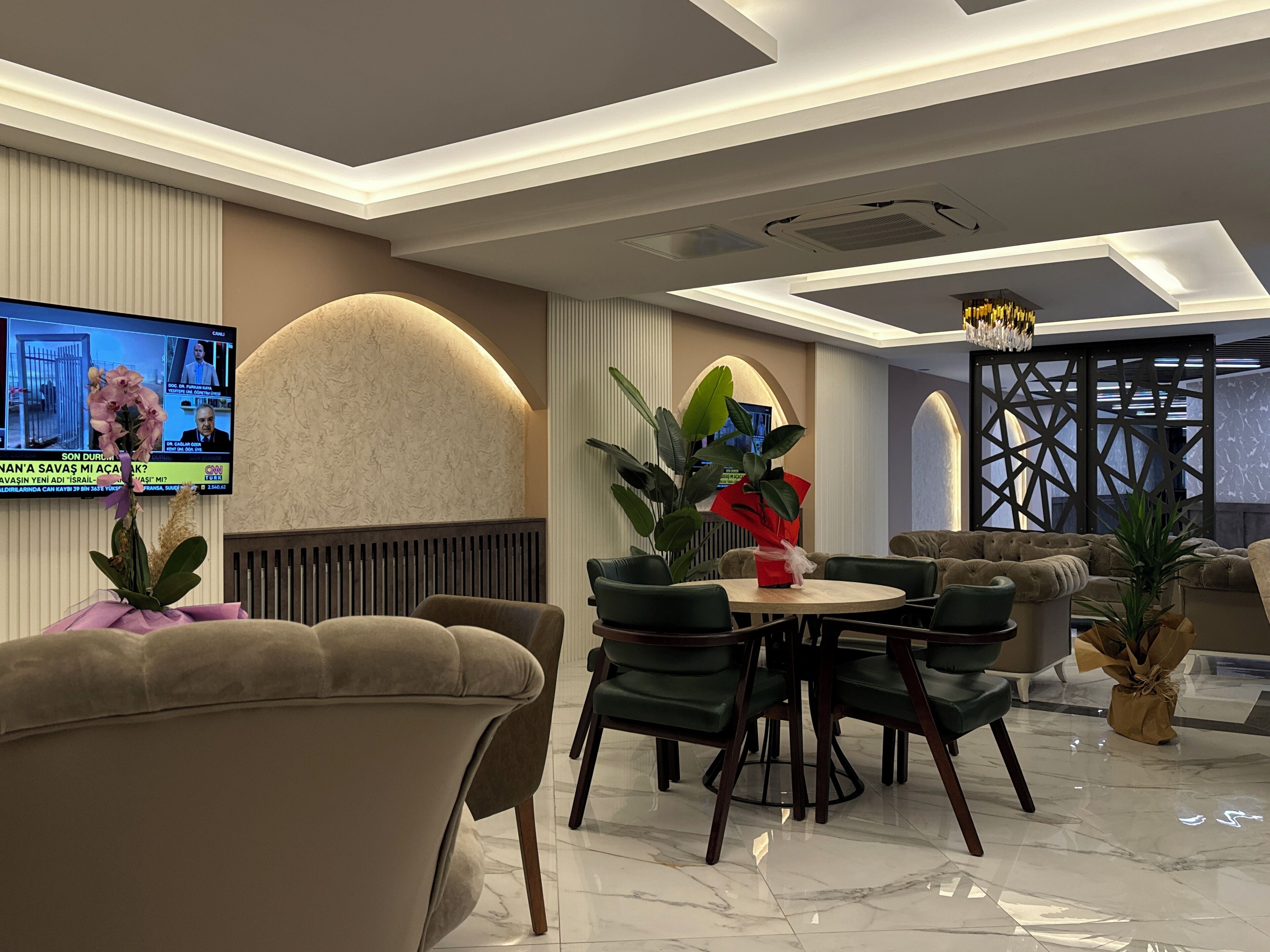 A modern lounge area with a TV displaying news, comfortable seating, and decorative plants.