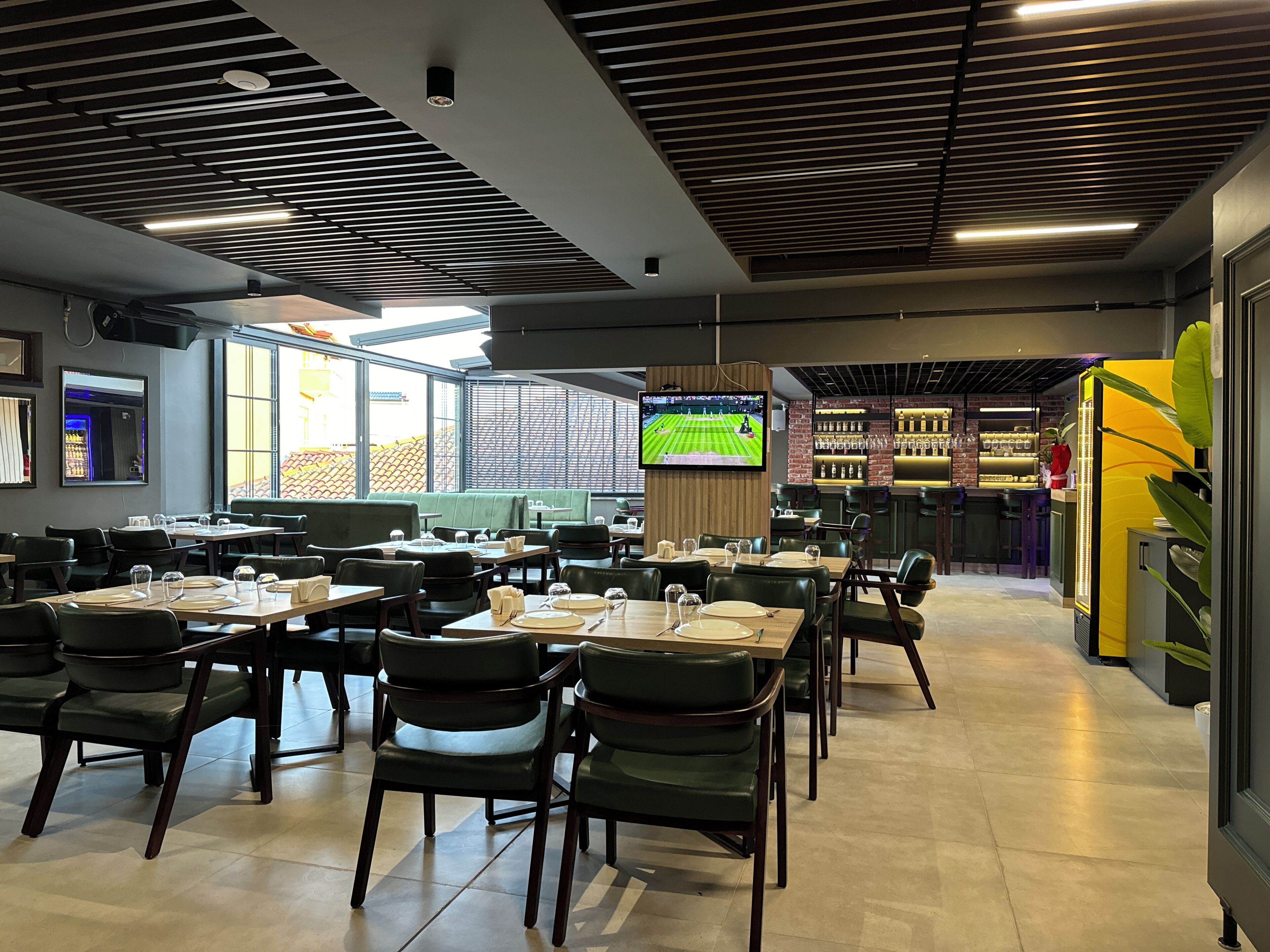 A modern restaurant with multiple dining tables and chairs, a bar area, and a large TV screen displaying a sports event.