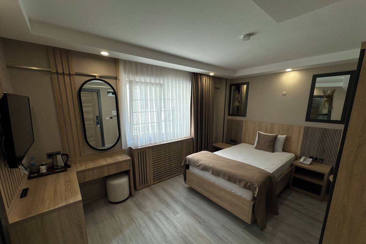 A modern hotel room with a large bed, a desk, a mirror, and a window with curtains.