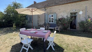 Un área de comedor al aire libre con una mesa, sillas, un paraguas grande y un edificio rústico al fondo.