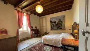 Un dormitorio con cama, escritorio, silla, radiador, ventana con cortinas, un cuadro en la pared y techo de madera.