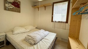 A bedroom with a bed, a nightstand, a wooden shelf, a window with a lace curtain, and a wall-mounted painting. - Cozy 1-Bedroom Apt in Bernex - Ski-In/Ski-Out, WiFi, Pet-Friendly, Near Evian & Lake Geneva (Bernex)