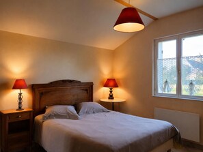 Un dormitorio con cabecera de madera, dos mesitas de noche con lámparas, una ventana con cortinas de encaje y un radiador.