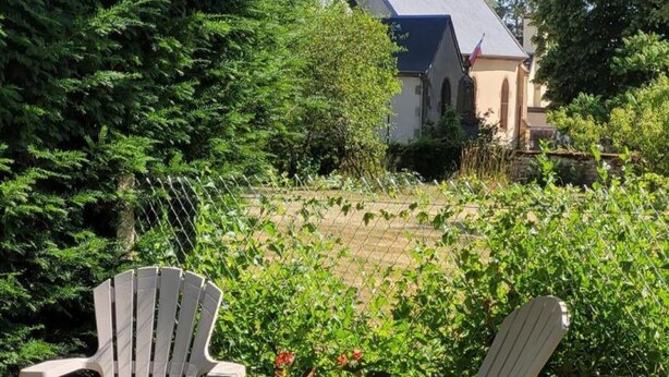 Dos sillas Adirondack blancas mirando hacia un jardín con una cerca, un edificio de techo inclinado y abundante vegetación.