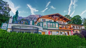 Un hotel de estilo alpino tradicional, con balcones adornados con macetas de flores, un letrero llamativo y vistas a las montañas al fondo.
