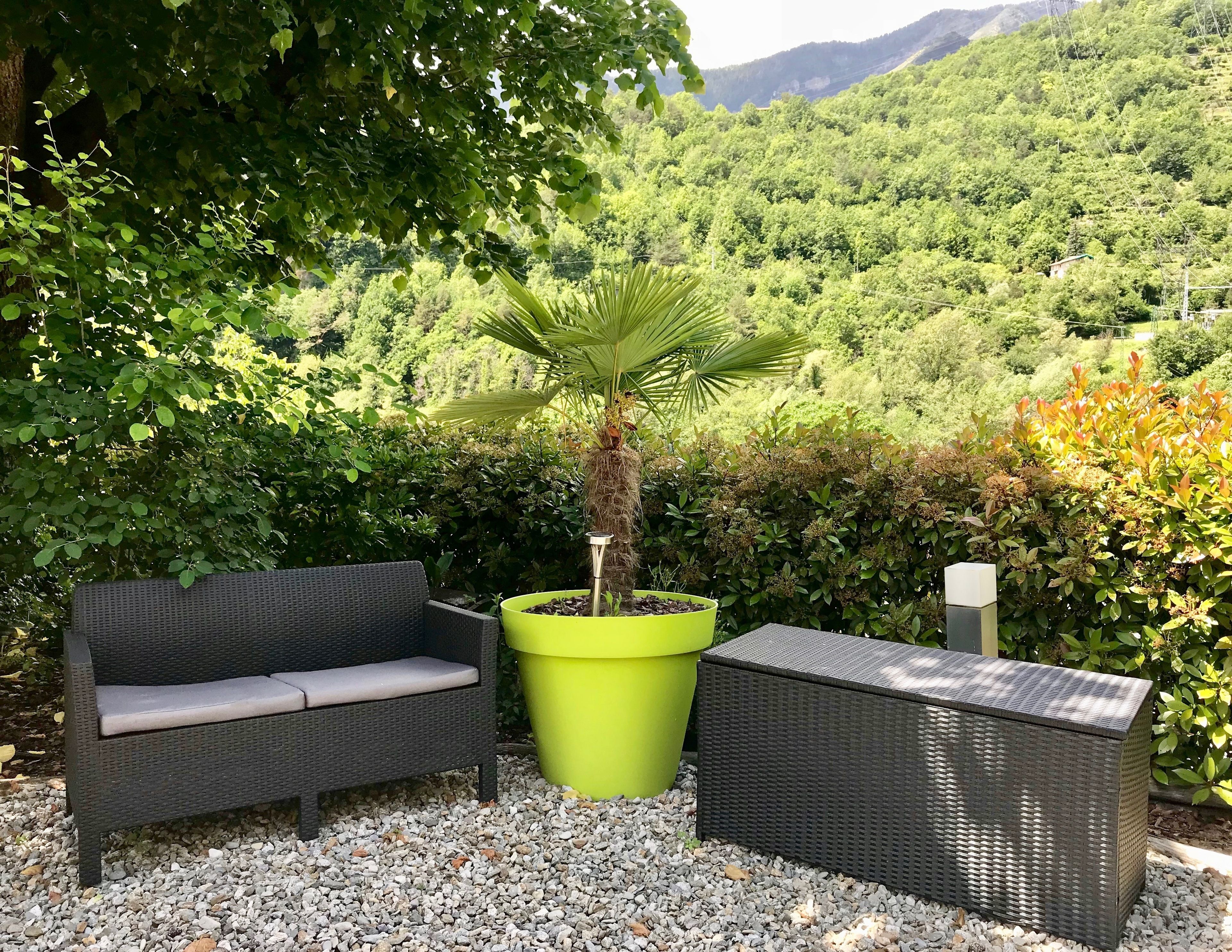 Un canapé en osier et un pouf assorti sont posés sur un sol de gravier, entourés d’une végétation luxuriante et avec une chaîne de montagnes en toile de fond.