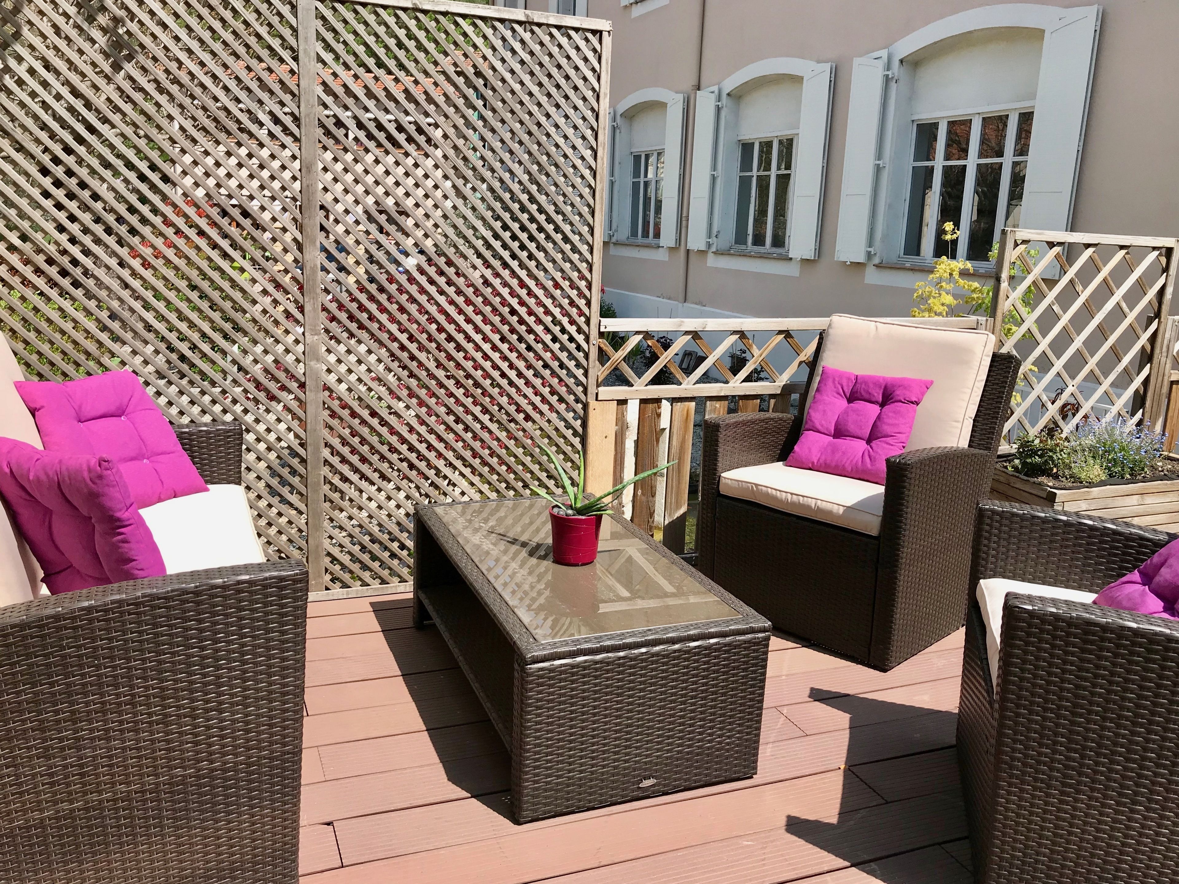Un espacio de patio al aire libre con mobiliario de mimbre, una mesa de vidrio y un fondo de celosía de madera.