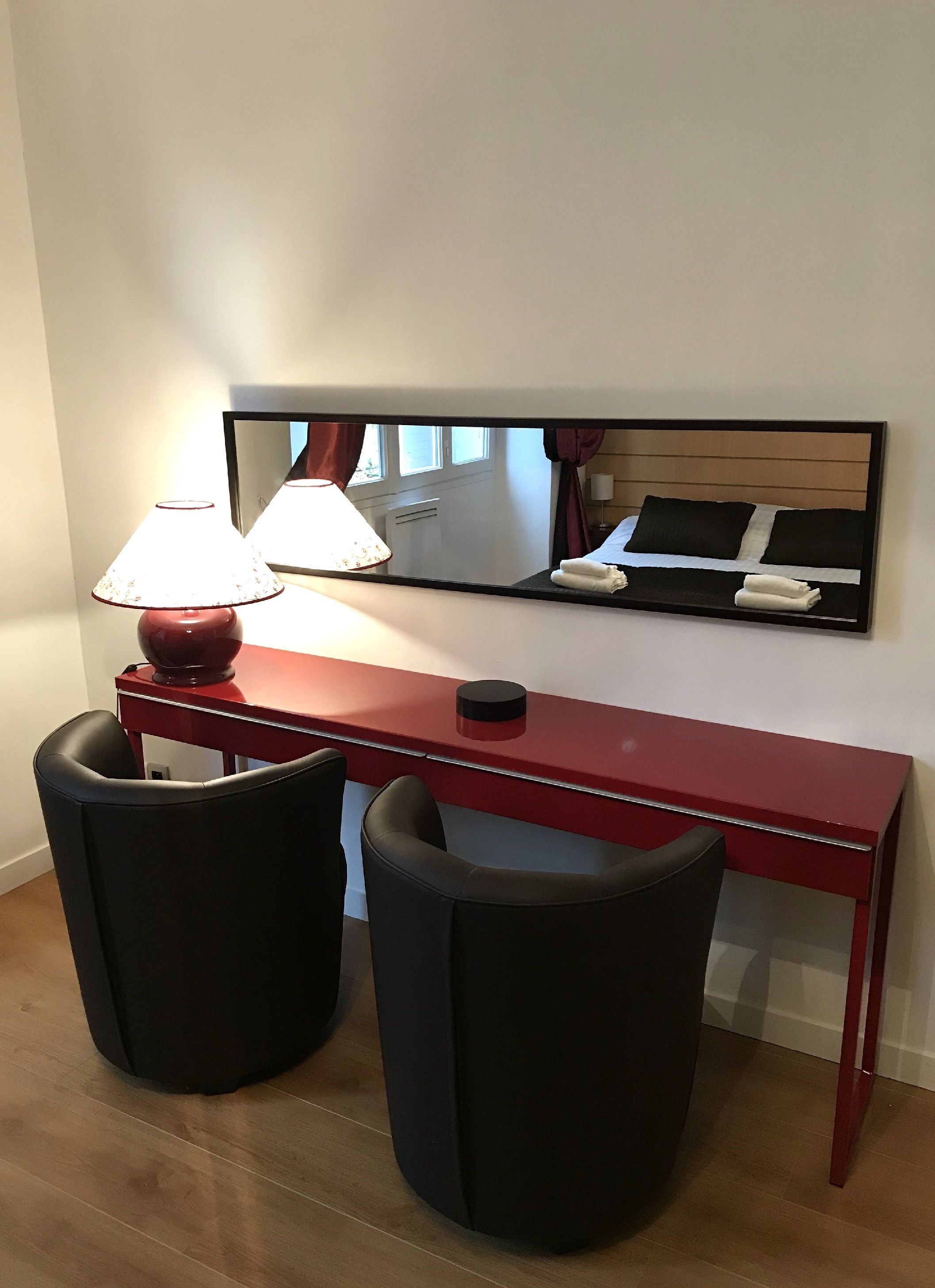 Een hotelkamer met een rode bureau, twee zwarte stoelen, een lamp en een spiegel die het bed en het raam weerspiegelt.