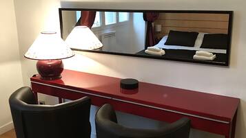 Een hotelkamer met een rode bureau, twee zwarte stoelen, een lamp en een spiegel die het bed en het raam weerspiegelt.