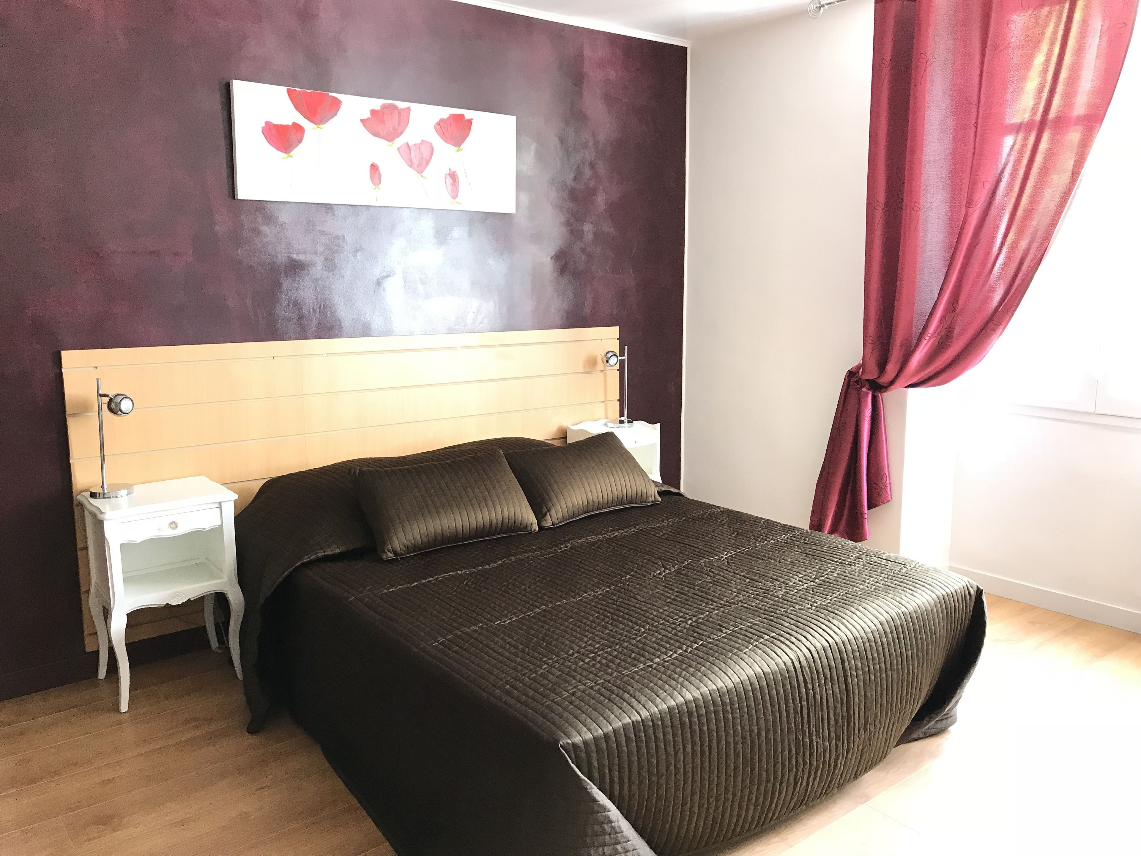 A bedroom with a wooden headboard, a bed with a dark quilted bedspread, a nightstand with a lamp, and a wall art piece above the bed.