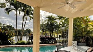 A poolside area with a table and chairs, a ceiling fan, and palm trees in the background. - Pristine Home in Golf Community 3 Bedroom/3.5 Bathroom (West Palm Beach)