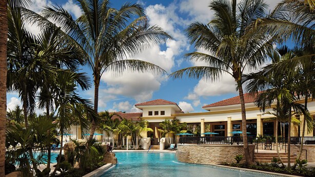 A resort pool area with palm trees, a clear sky, and a building with a red roof. - Pristine Home in Golf Community 3 Bedroom/3.5 Bathroom (West Palm Beach)