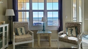 Una stanza con due poltrone bianche con cuscini a cavalluccio marino, un tavolino con una lampada e vista sul mare attraverso ampie finestre.