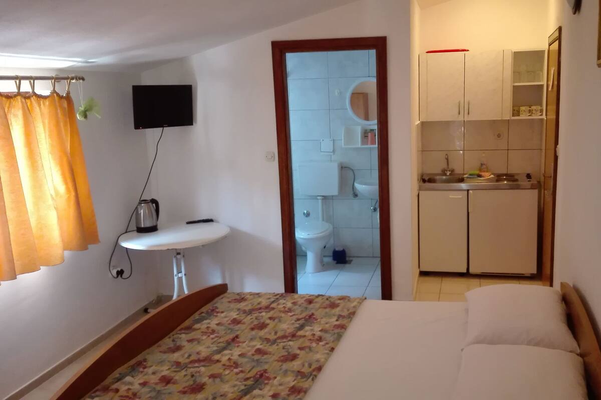 Ein Schlafzimmer mit einem Bett, einem Nachttisch mit Wasserkocher, einem kleinen Fenster mit Vorhängen und Blick auf ein Badezimmer und eine Küchenzeile.