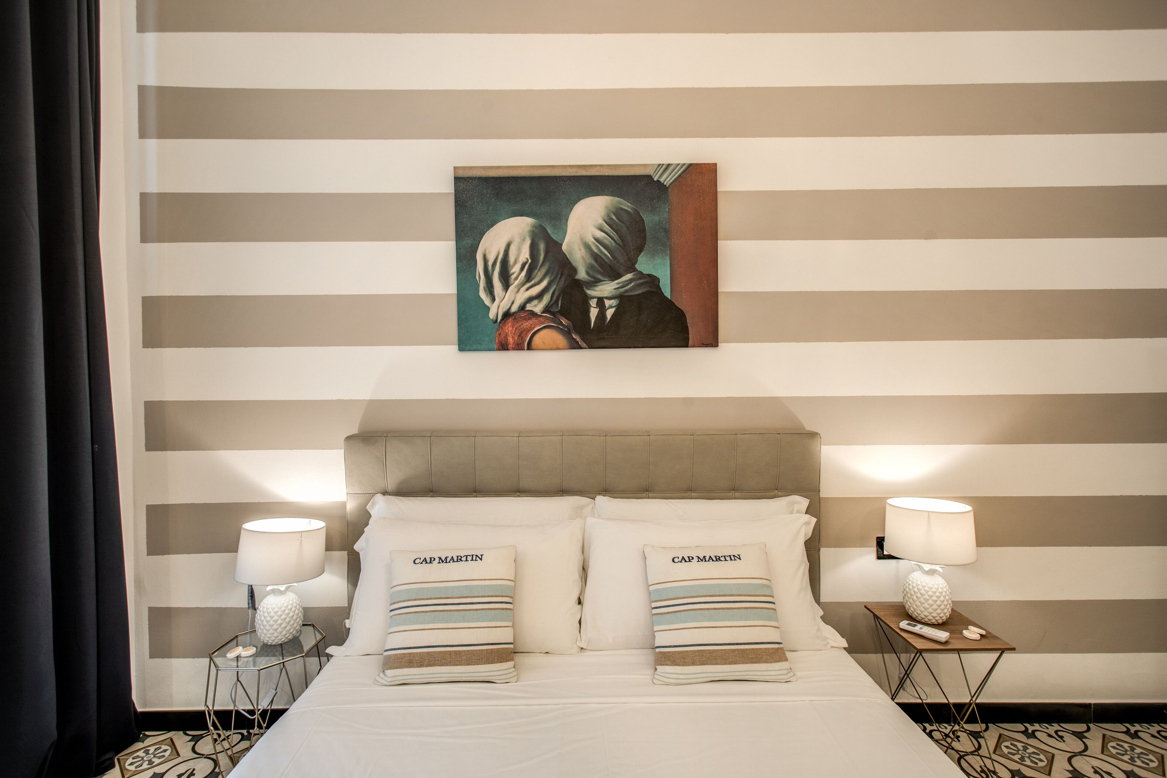 Quarto de hotel com parede listrada, um quadro com duas pessoas, duas luminárias e almofadas com a inscrição 'CAP MARTIN'.