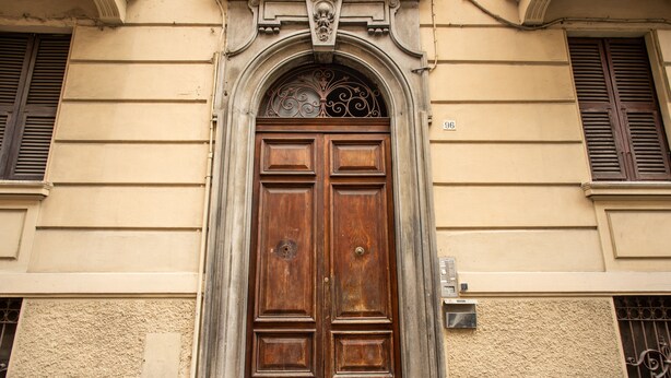 Puerta doble de madera con un arco decorativo encima, en un edificio con paredes color beige y una ventana con contraventanas.