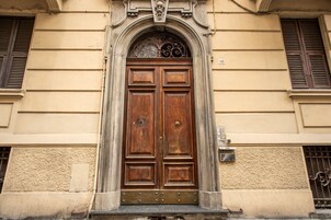 Puerta doble de madera con un arco decorativo encima, en un edificio con paredes color beige y una ventana con contraventanas.
