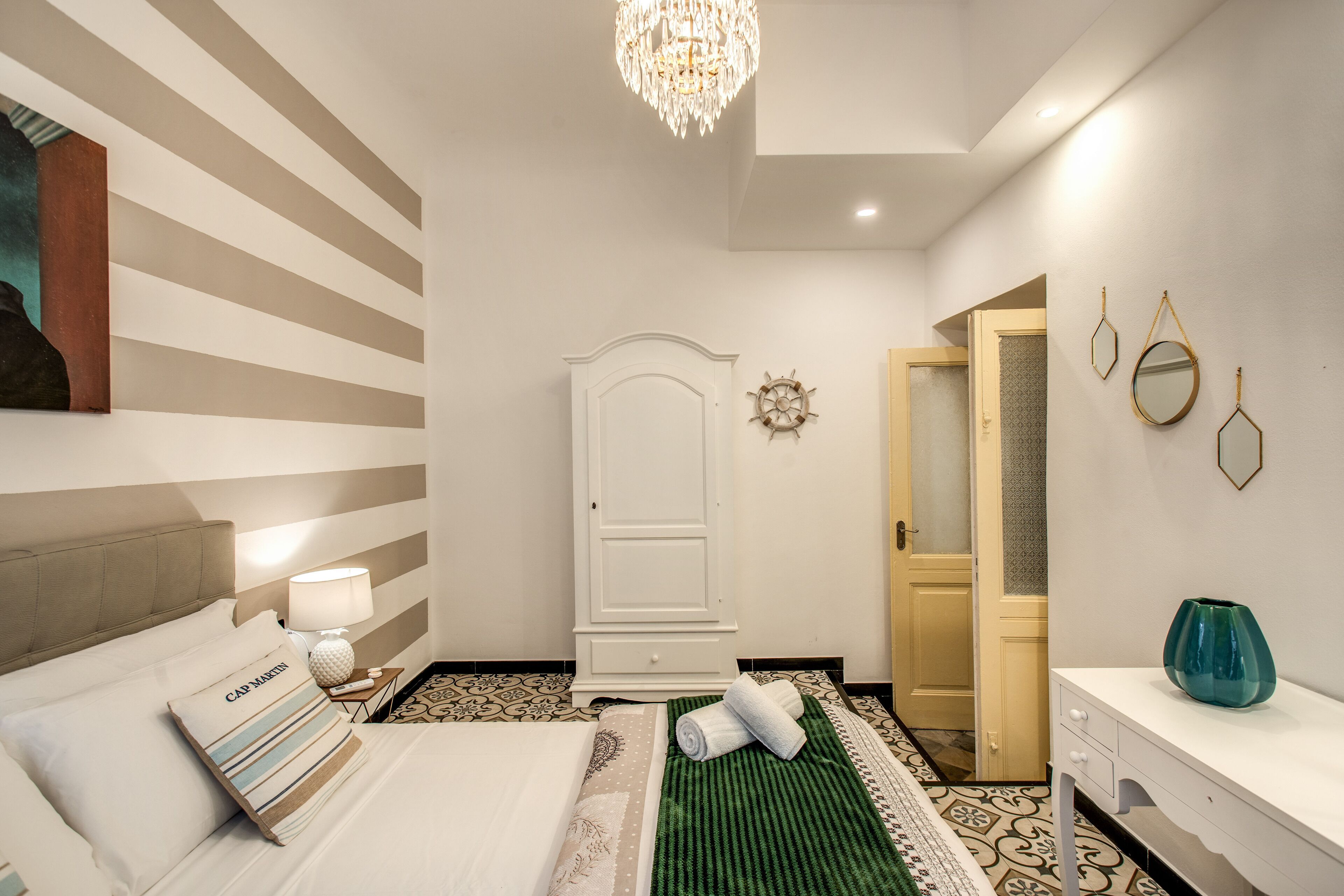 Una habitación de hotel con cama, mesita de noche con lámpara, cómoda, espejo y puerta que da a otra habitación.