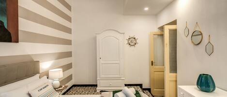 Una habitación de hotel con cama, mesita de noche con lámpara, cómoda, espejo y puerta que da a otra habitación.