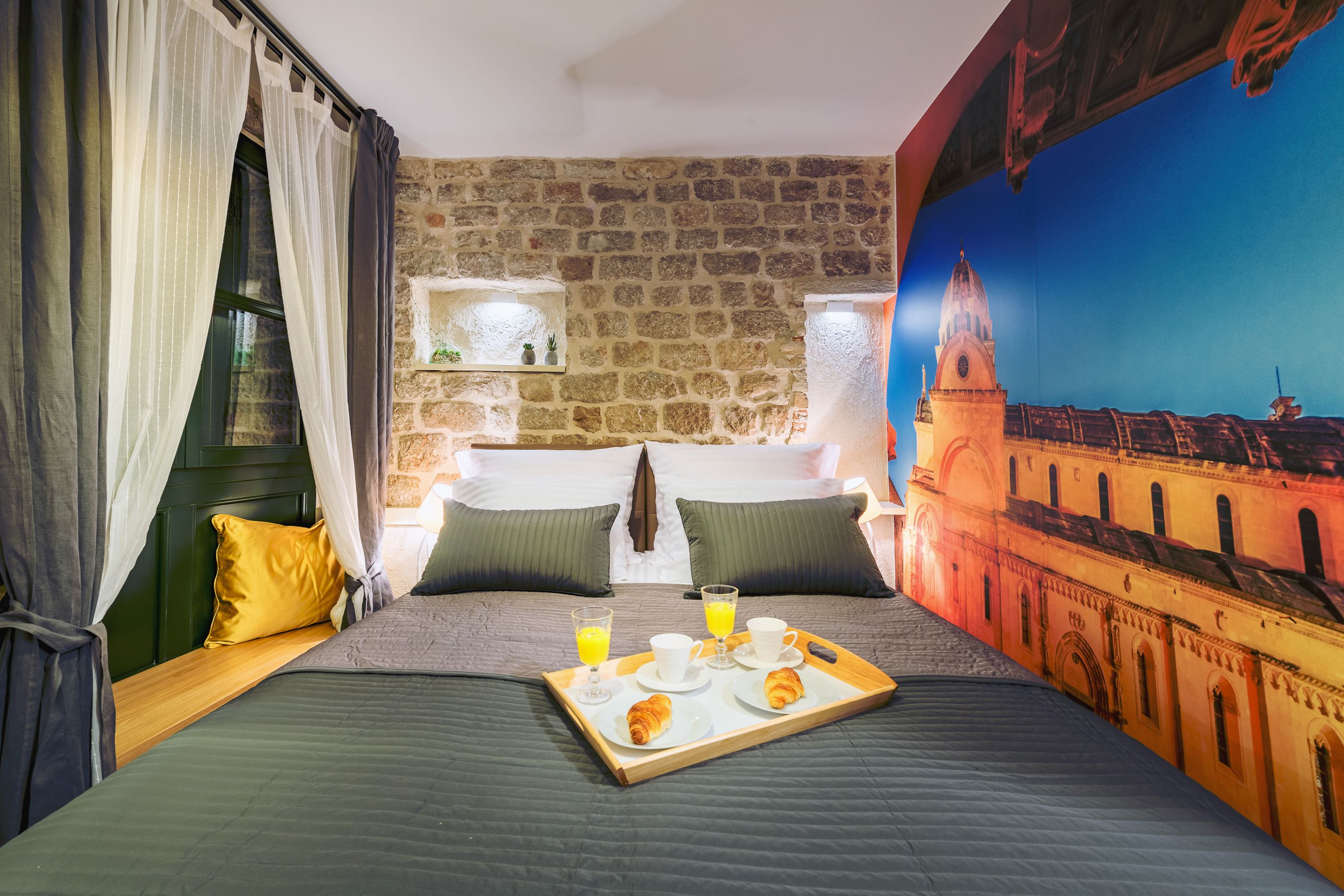 Une chambre d’hôtel avec un mur en pierre, un lit avec une literie verte, un plateau de petit-déjeuner avec des croissants et une fresque représentant un bâtiment.