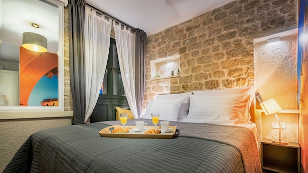 Une chambre d’hôtel chaleureuse avec un mur en pierre, un lit recouvert de draps blancs, une table de chevet avec une lampe et un plateau de petit-déjeuner comprenant des croissants et du jus d’orange.