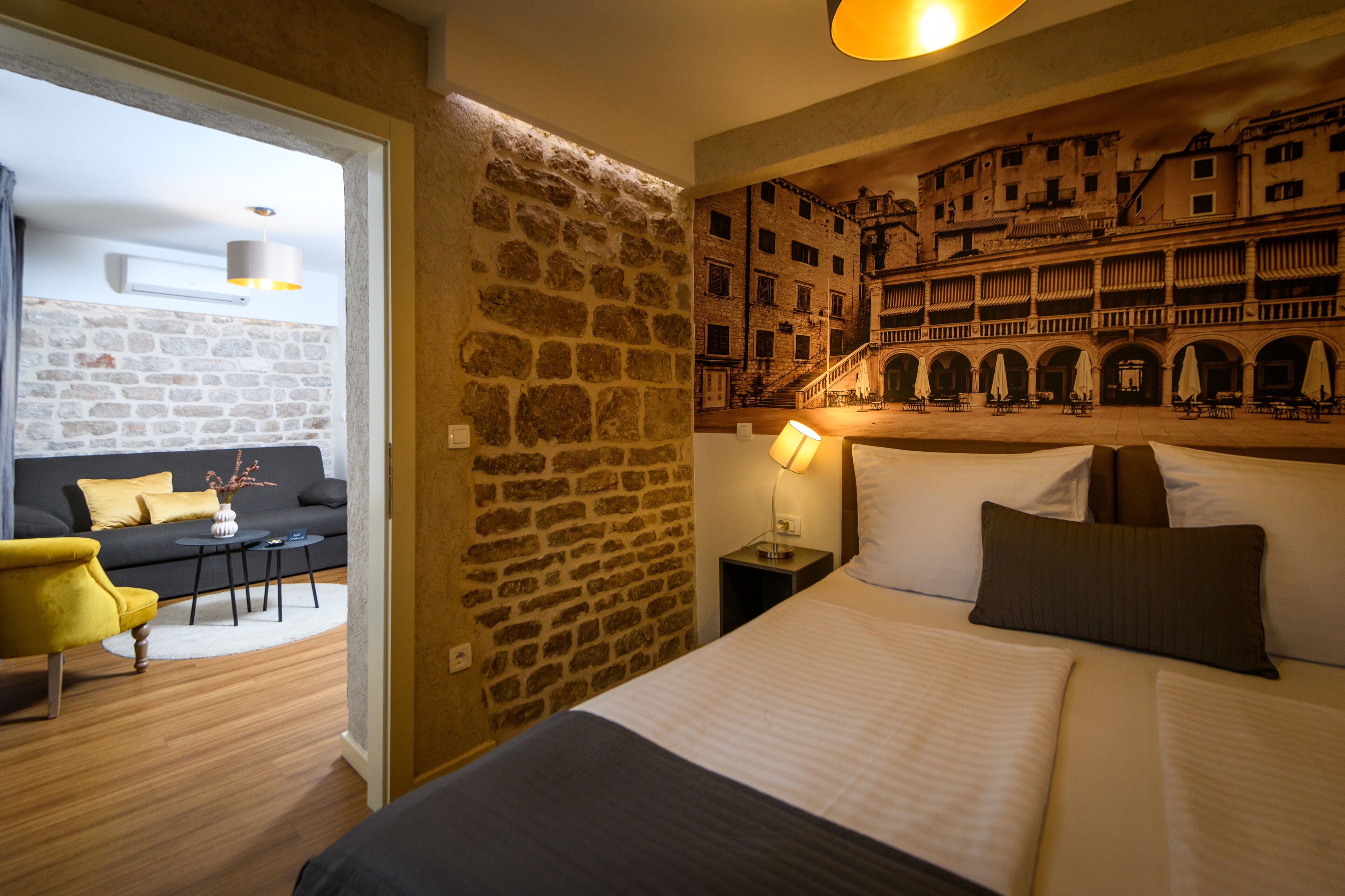 Une chambre d’hôtel avec un mur en pierre, un lit avec des oreillers, une table de chevet avec une lampe, un canapé avec des coussins et une vue sur un immeuble par la fenêtre.