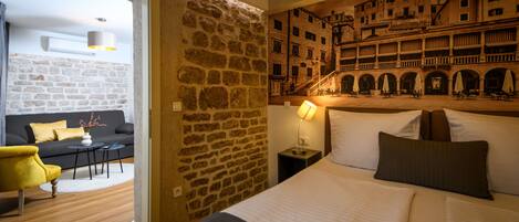Une chambre d’hôtel avec un mur en pierre, un lit avec des oreillers, une table de chevet avec une lampe, un canapé avec des coussins et une vue sur un immeuble par la fenêtre.