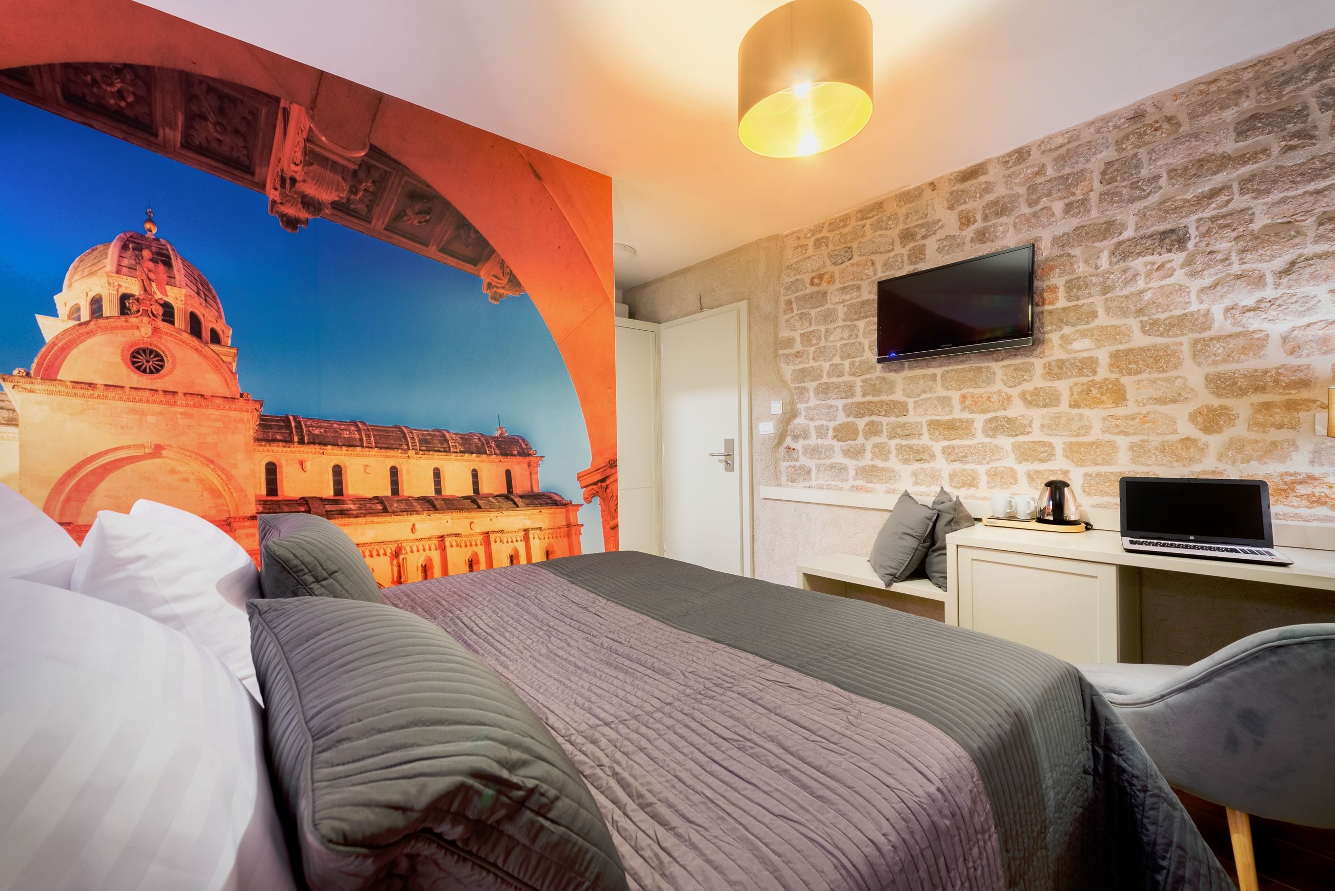 A hotel room with a large bed, a desk with a laptop, a TV mounted on a stone wall, and a mural of a historical building.