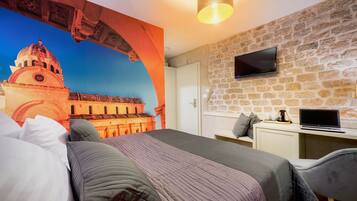 Una habitación de hotel con una cama grande, un escritorio con una computadora portátil, un televisor montado en una pared de piedra y un mural de un edificio histórico.