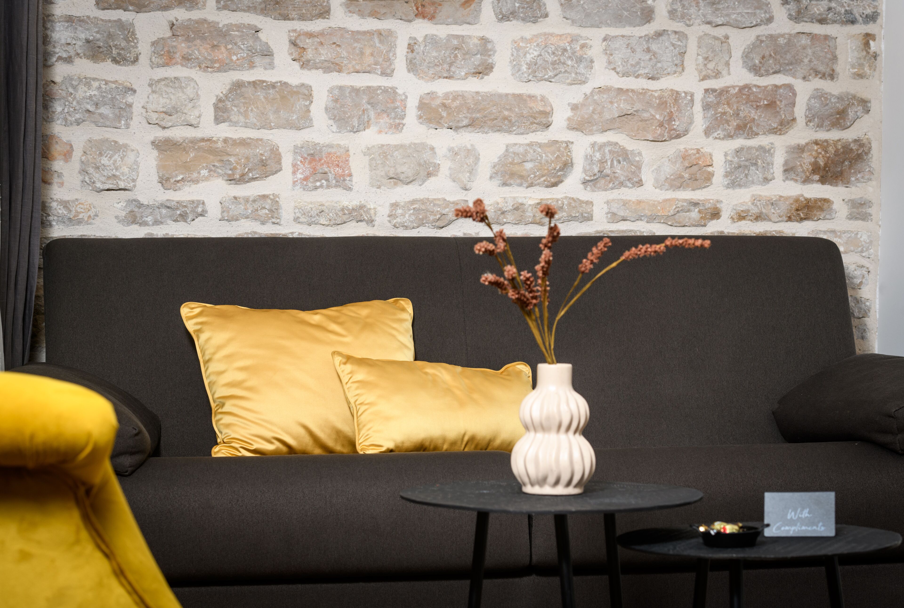 A modern room with a stone wall, a dark sofa with yellow cushions, a white vase with dried flowers, and a small table with a sign.