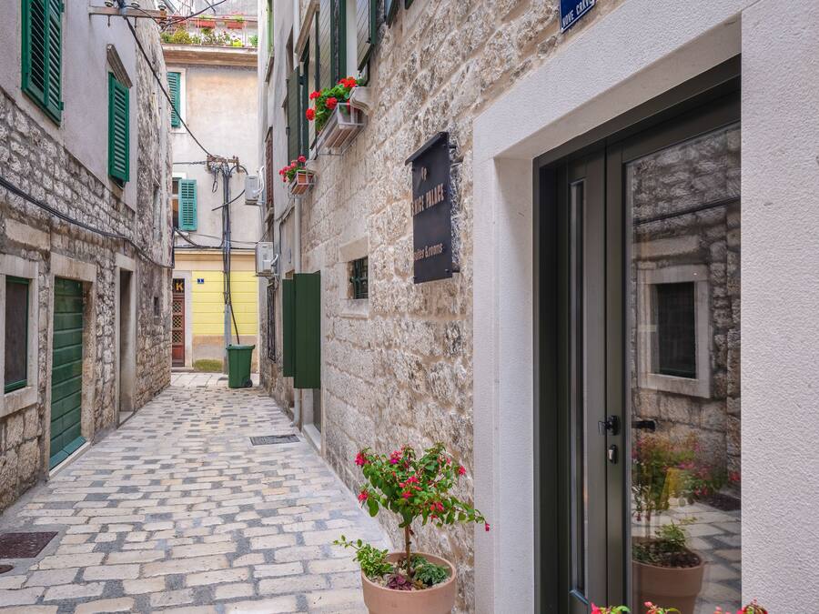 A narrow cobblestone street with stone buildings, green shutters, and a potted plant with flowers.