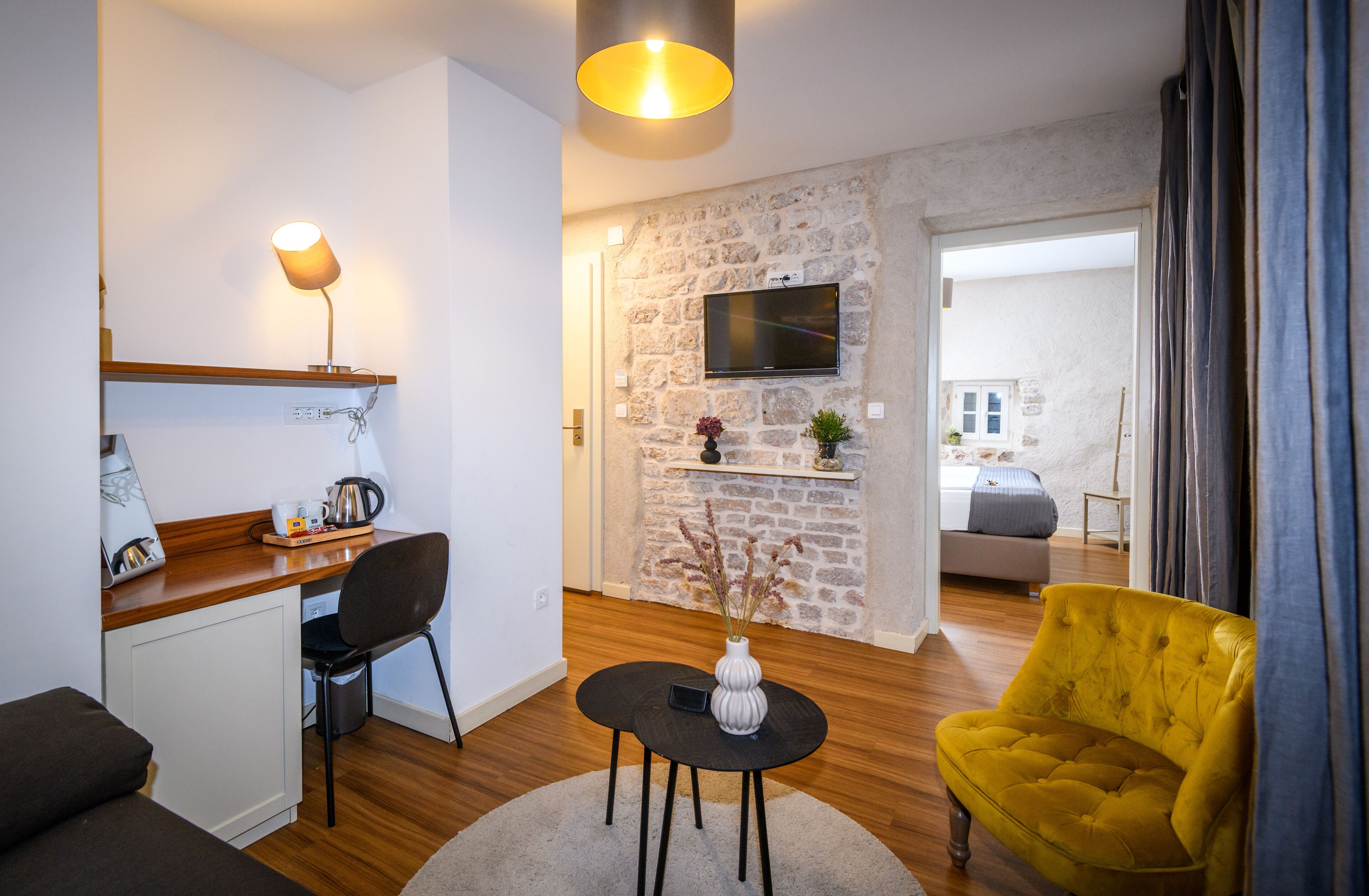 A modern living room with a stone wall, a flat-screen TV mounted above a fireplace, a yellow armchair, and a dining area with a wooden table and chairs.