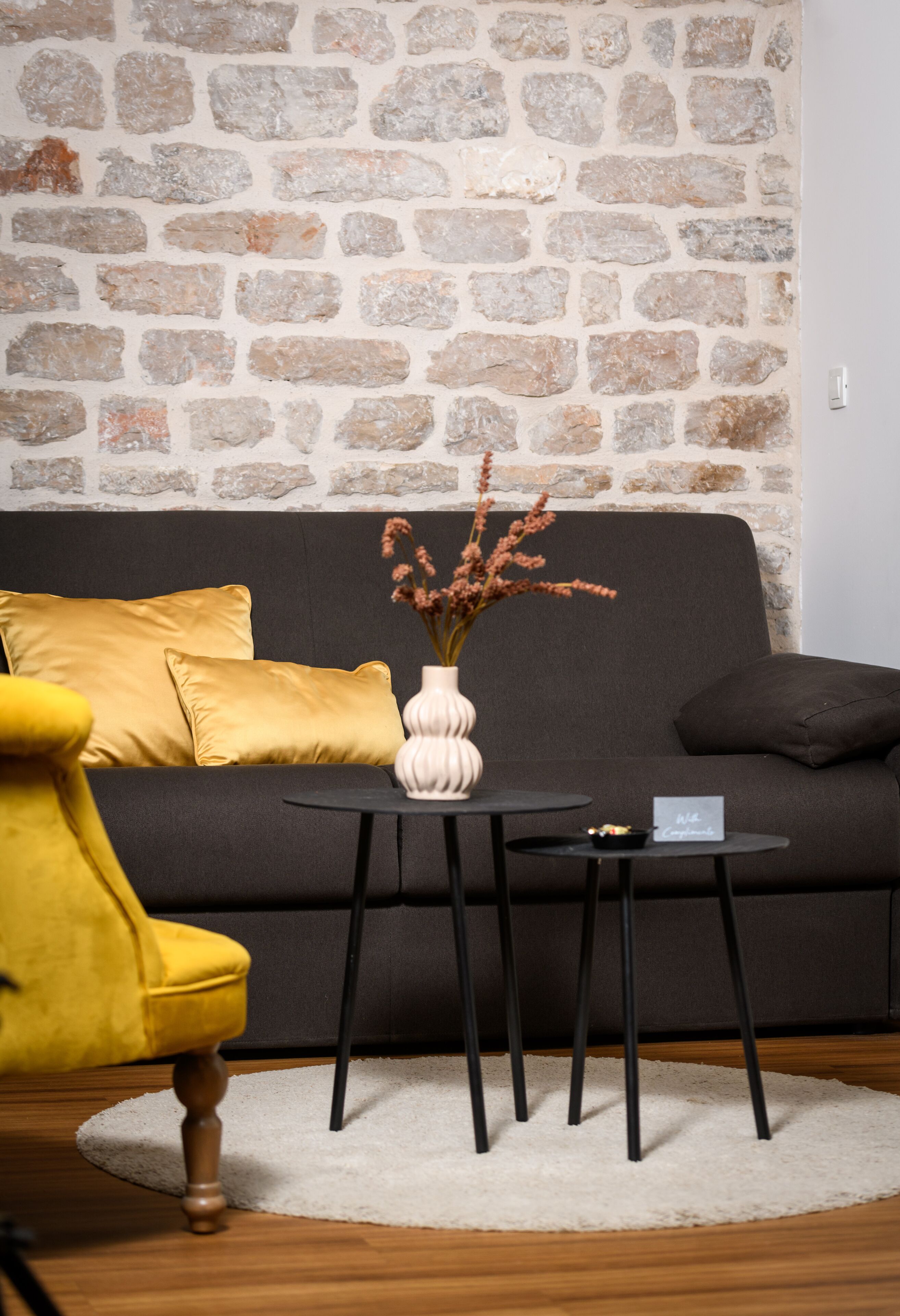 A room with a stone wall, a dark sofa, a yellow armchair, a small round table with a vase of dried flowers, and a white rug.