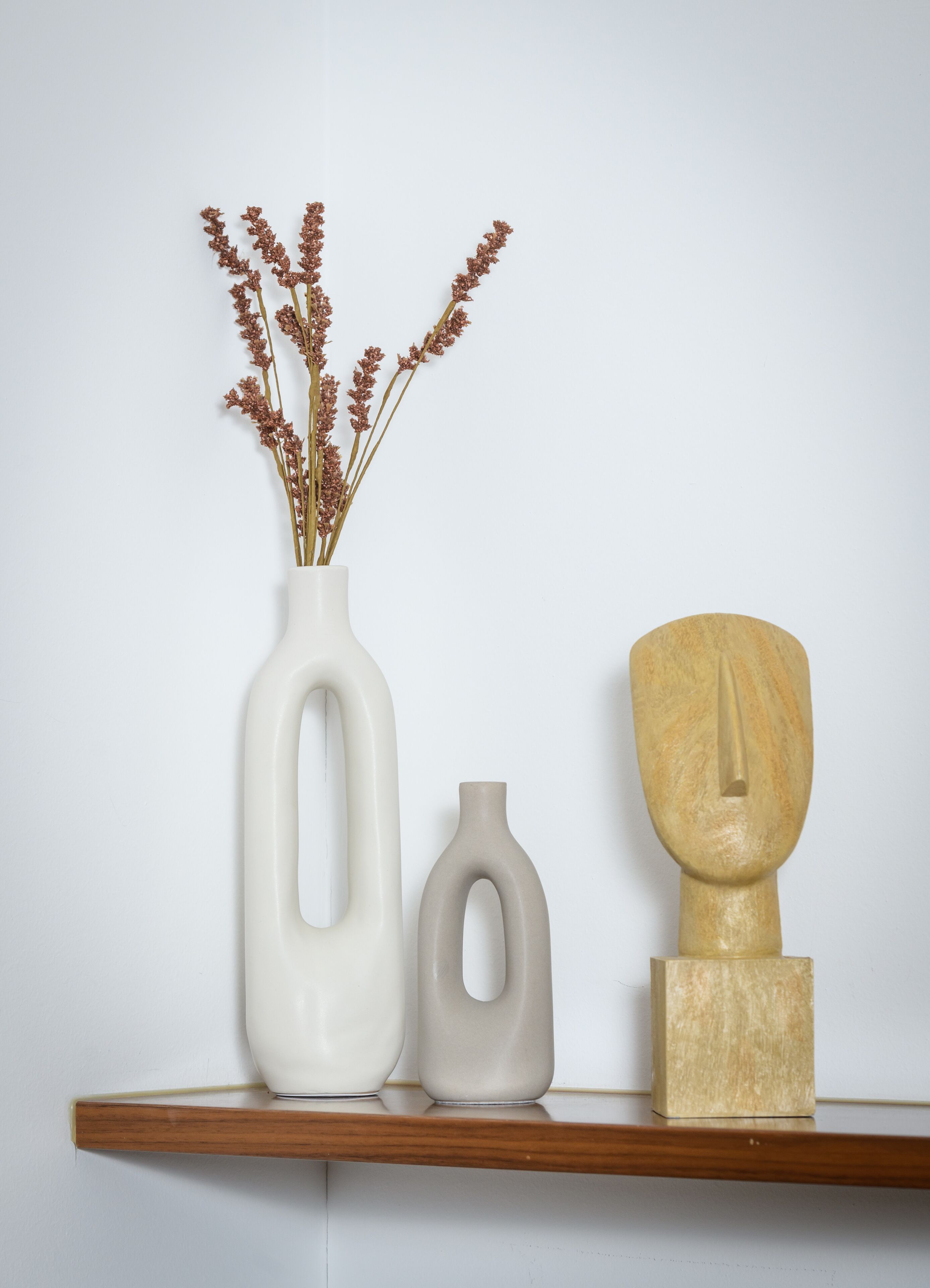 A shelf with three vases: a tall white vase with dried flowers, a smaller white vase, and a wooden sculpture.