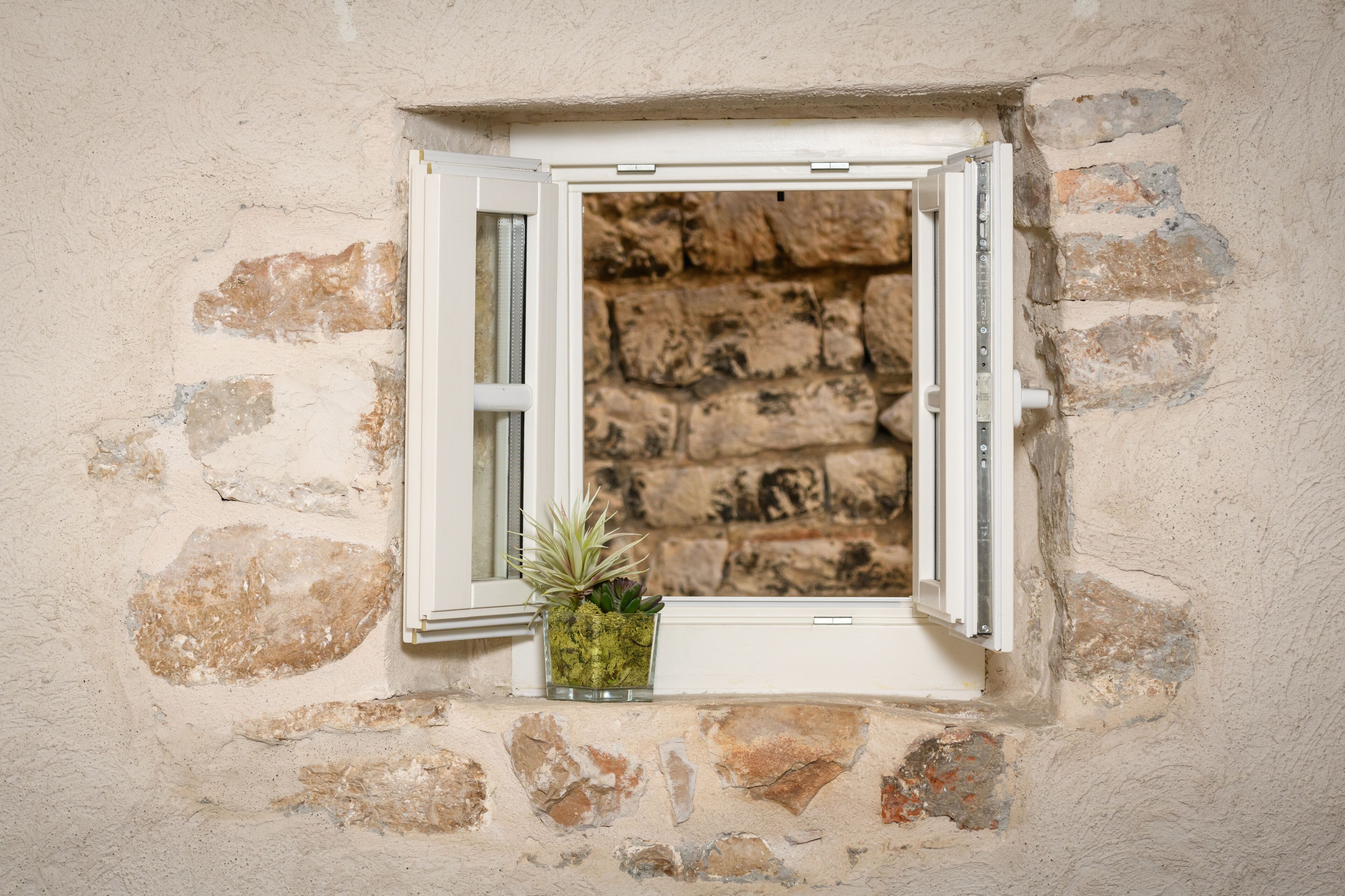 A small window with an open white shutter, a potted plant, and a stone wall.