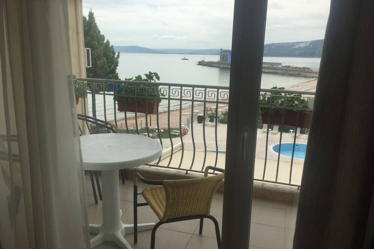 Un balcón con una mesa blanca y sillas de mimbre, con vista a una piscina y una playa.