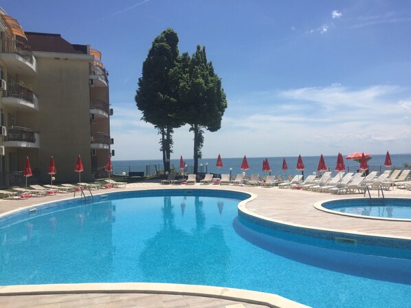 A pool area with lounge chairs and umbrellas, a clear sky, and a view of the sea.
