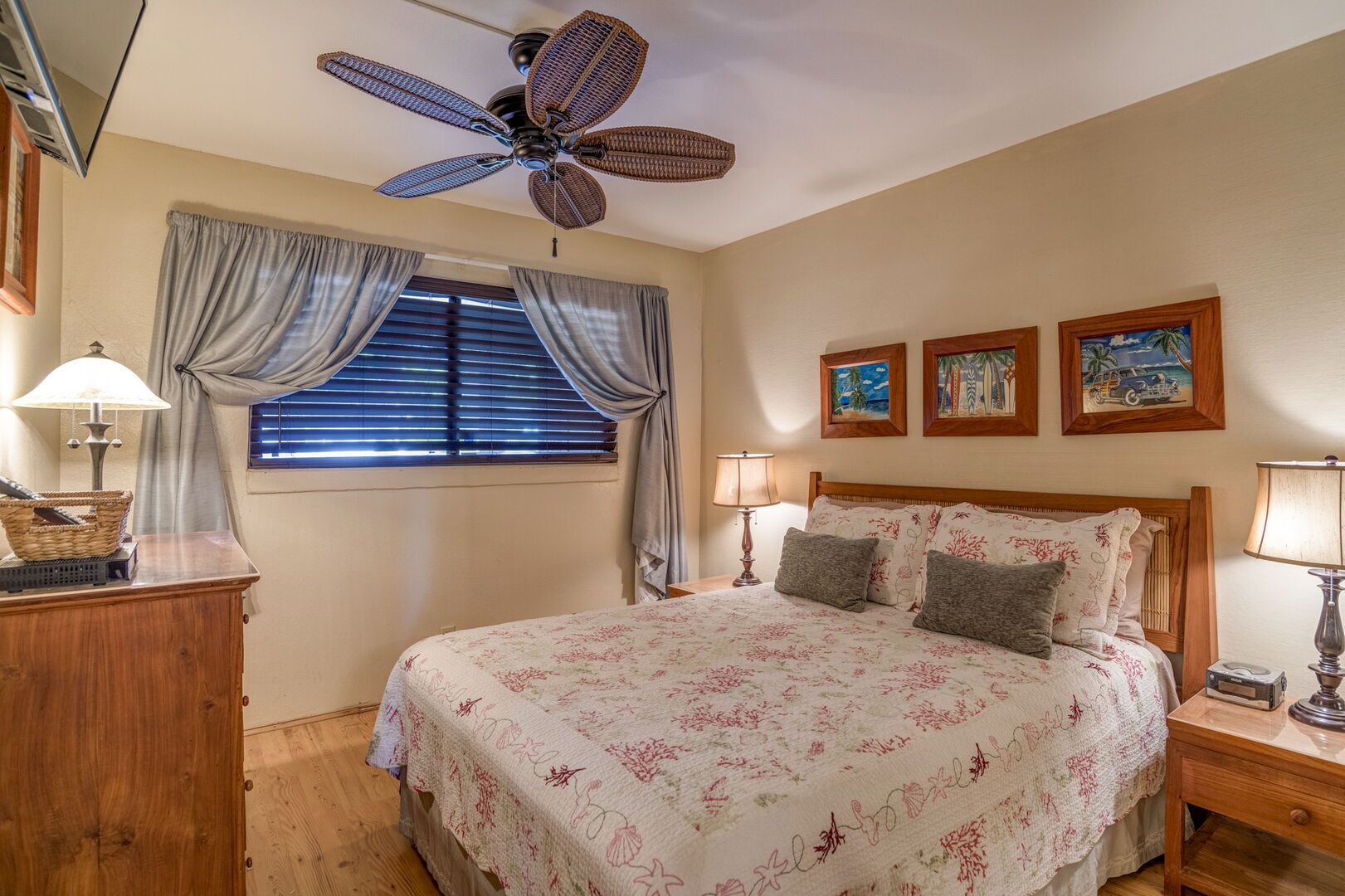 A bedroom with a bed, a ceiling fan, a window with curtains, a bedside table, and wall art.