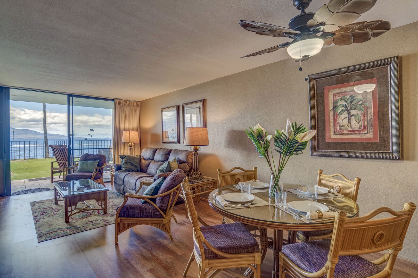 A spacious living room with a large window overlooking a scenic view, a dining area with a round table, and a ceiling fan.