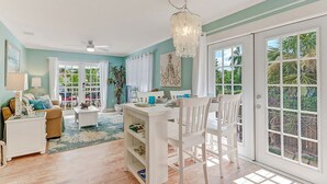 A spacious living room with a dining area, large windows, and a view of greenery outside. - Classic Beach-Style Cottage (Naples)