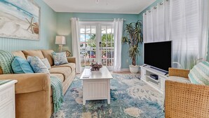 A living room with a beige sofa, a white coffee table, a flat-screen TV, and a large window with curtains. - Classic Beach-Style Cottage (Naples)