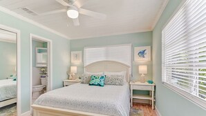 A bedroom with a bed, two bedside tables, a ceiling fan, a window with blinds, and a mirror. - Classic Beach-Style Cottage (Naples)