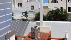 Une table et des chaises en bois, placées sous une pergola blanche, offrent une vue sur des bâtiments blancs et un ciel bleu dégagé.